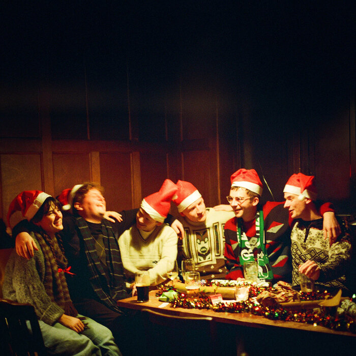 a group of people sitting at what appears to be a table at a pub, wearing santa hats and throwing their arms around each others' shoulders.