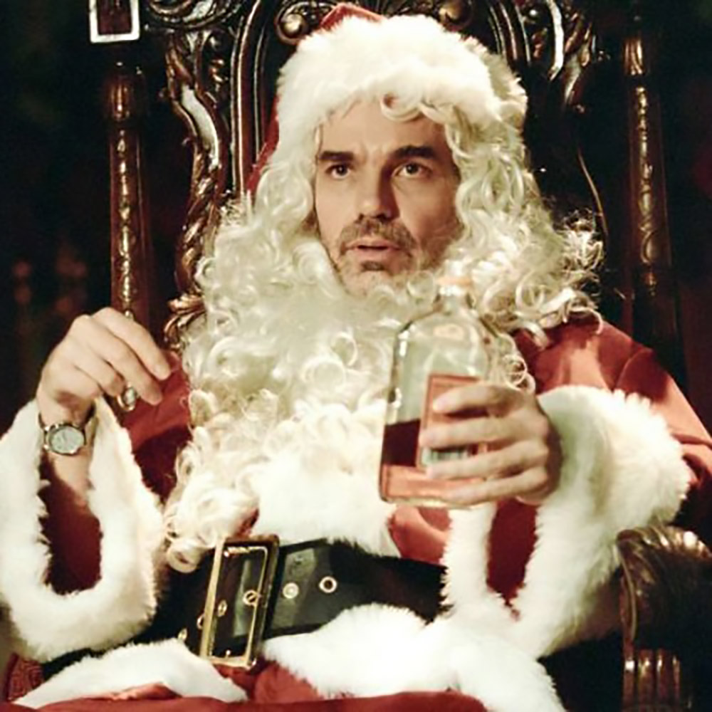 Billy Bob Thorton, dressed as Santa Clause from Bad Santa sitting in a chair holdng a drink.